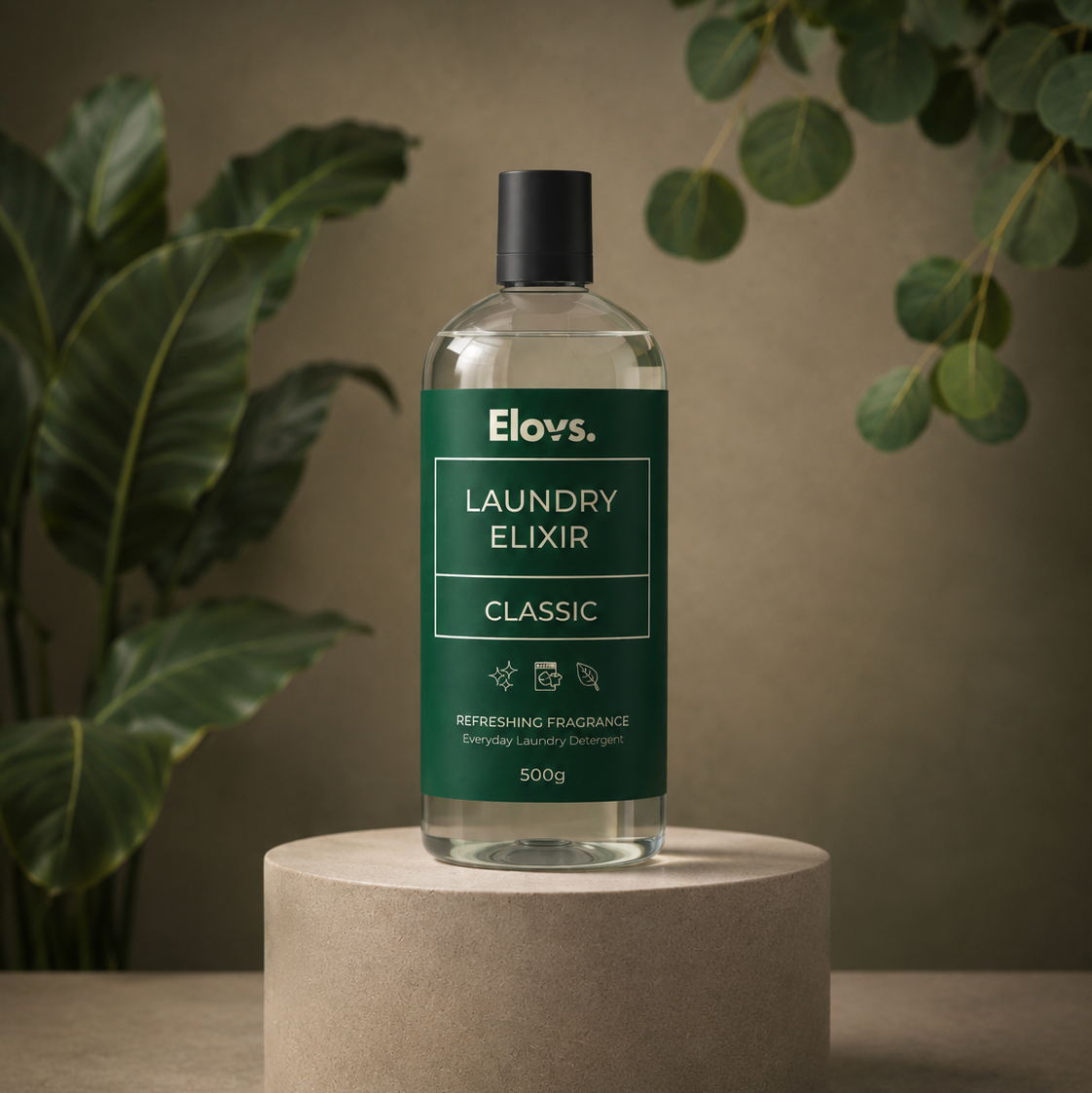 A bottle of Elov's Laundry Elixir Classic fragrance, 500g, displayed on a round concrete pedestal. The bottle is clear glass with a dark green label and a black cap, set against a warm brown background with soft-focus green plants.